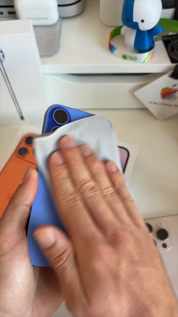 Hand using a grey microfiber cloth to wipe the dual camera lenses on the back of a blue iPhone